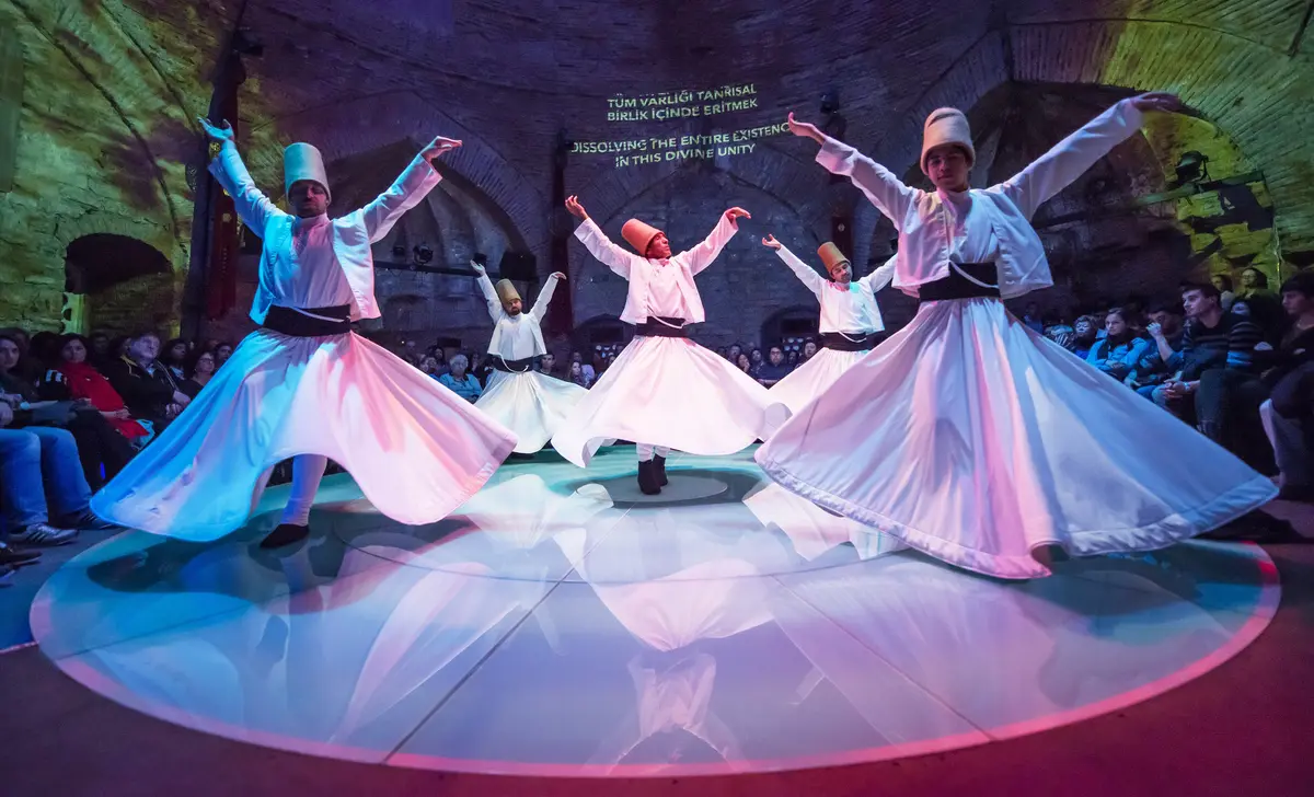 Four semazens in white skirts and tall brown hats turning with raised arms on a round floor, with seated spectators around them under a brick vault