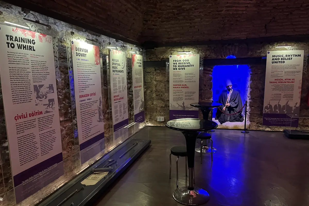 Information panels and glass display cases along a stone wall in a dim exhibition room, with a figure of a seated dervish playing a reed flute in a blue-lit alcove